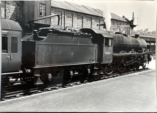 Jubilee No.45612 departs from an unidenited station with a passenger train. There's a goods warehouse behind