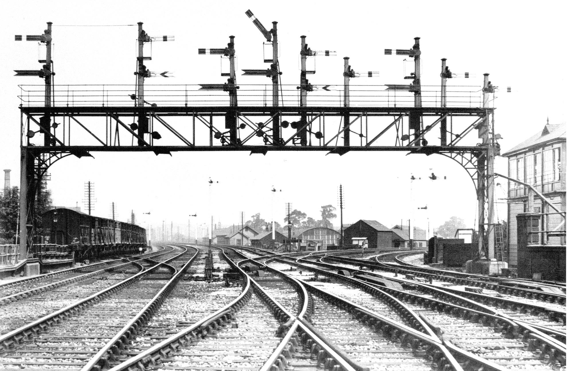 A large signal gantry spans four railway lines with a signal box just visible to the right. A colleciton of wooden buildings are visible ahead and, partially hiddne by a rake of cattle wagons, to the left as well.