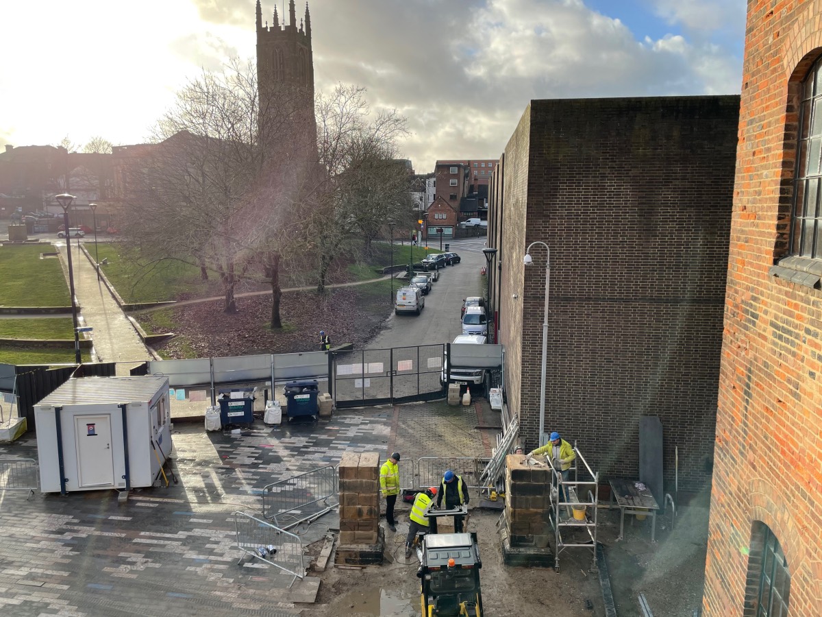 Viewed through a first floor window, workers are busy re-erecting two large stone pillars with Derby cathederal sillouetted by the low sun in the background
