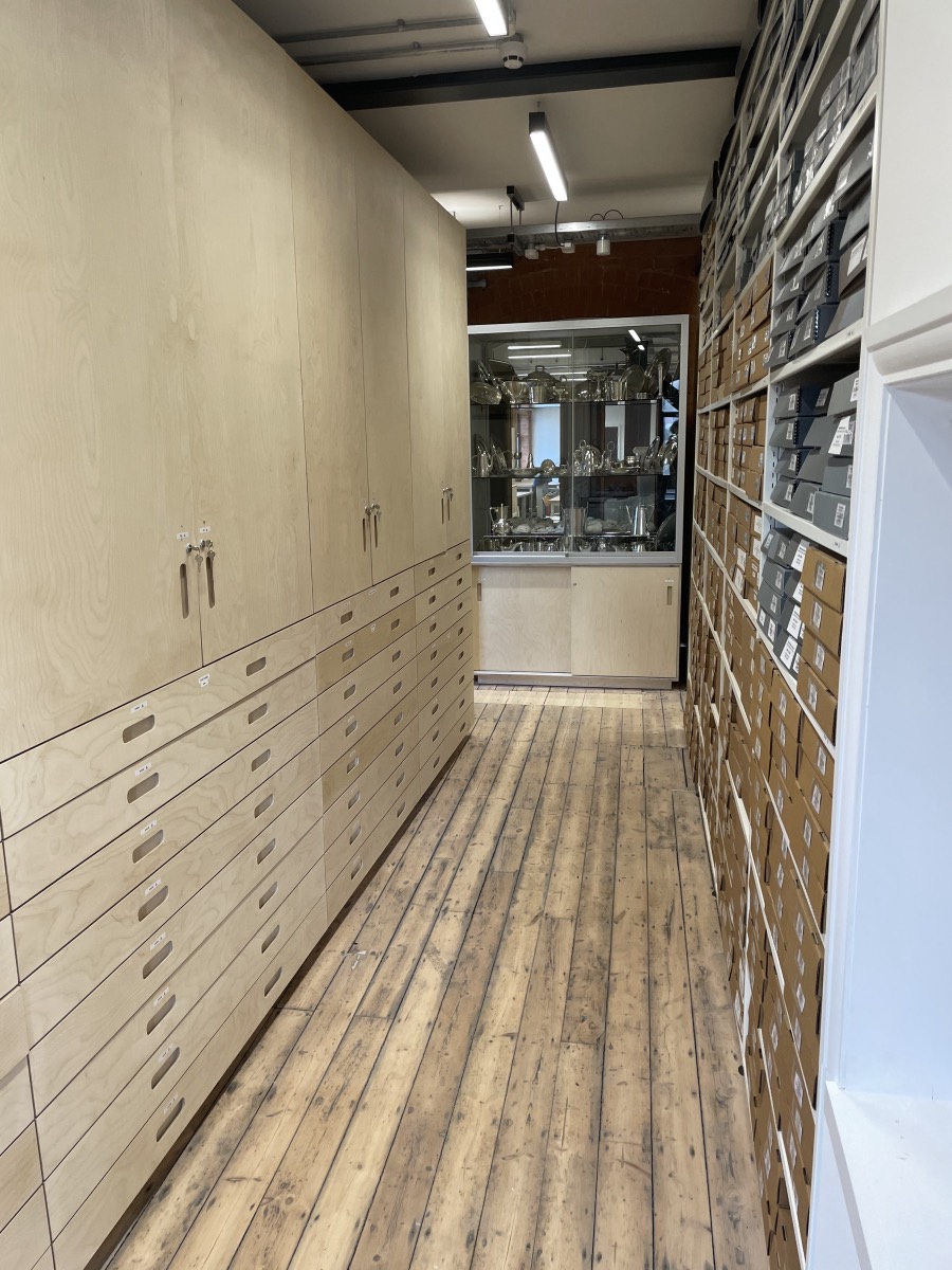 An aisle lined with steel shelves filled with archive boxes on the right and wooden cupboards and plan chest drawers below on the left