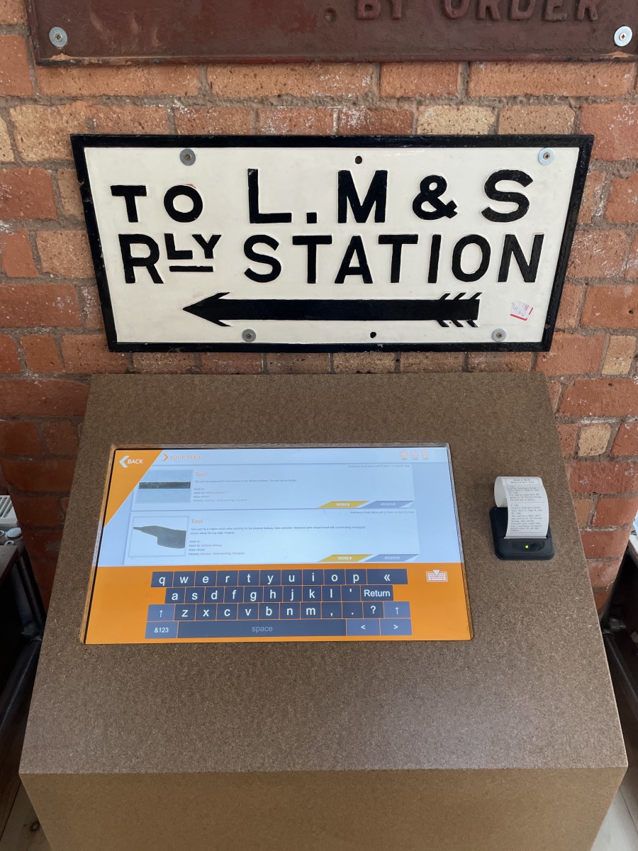 A computer workstation with a cast iron sign mounted on the wall above it reading 'To LMS Rly Station'