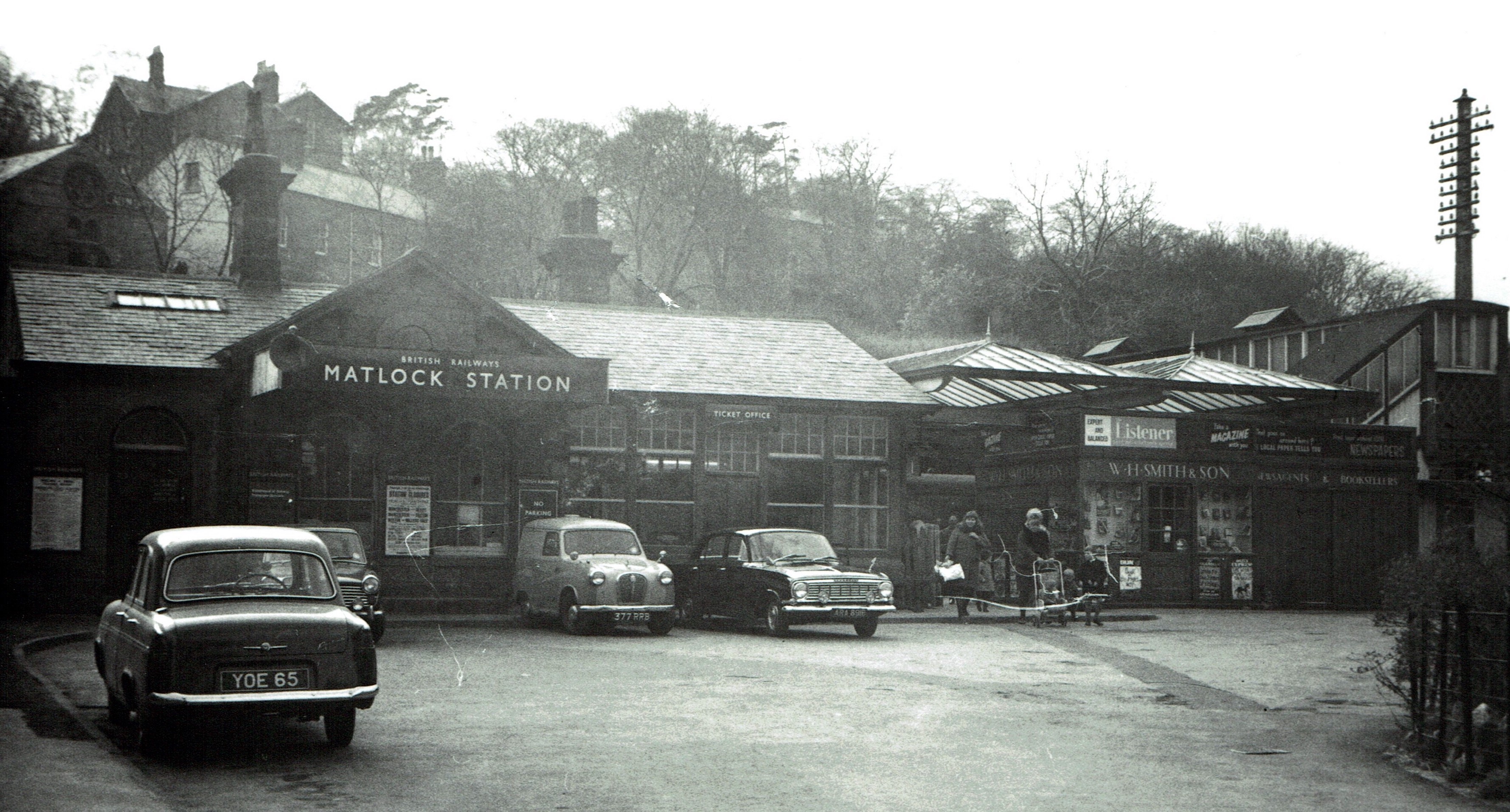 A black & white photograph taken in the mid 1960s of the frontage of Matock Railway station with cars parked in front of the booking office, a family with a push chair leaving the station and the ubiquitous W.H.Smith bookstall
