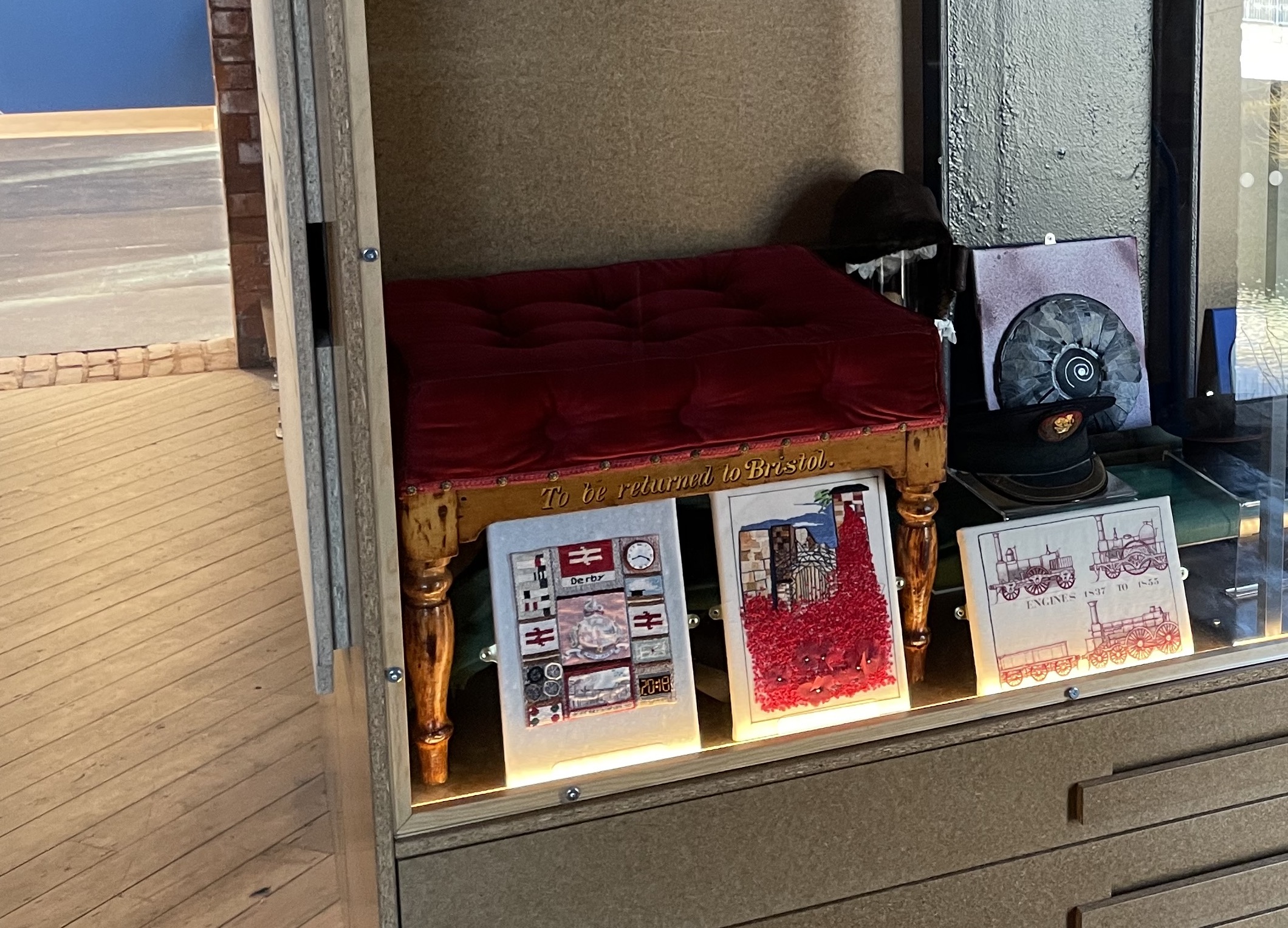 A low, highly varnished wooden stool with deep red upholstery continaed within a glass fronted display cabinet.