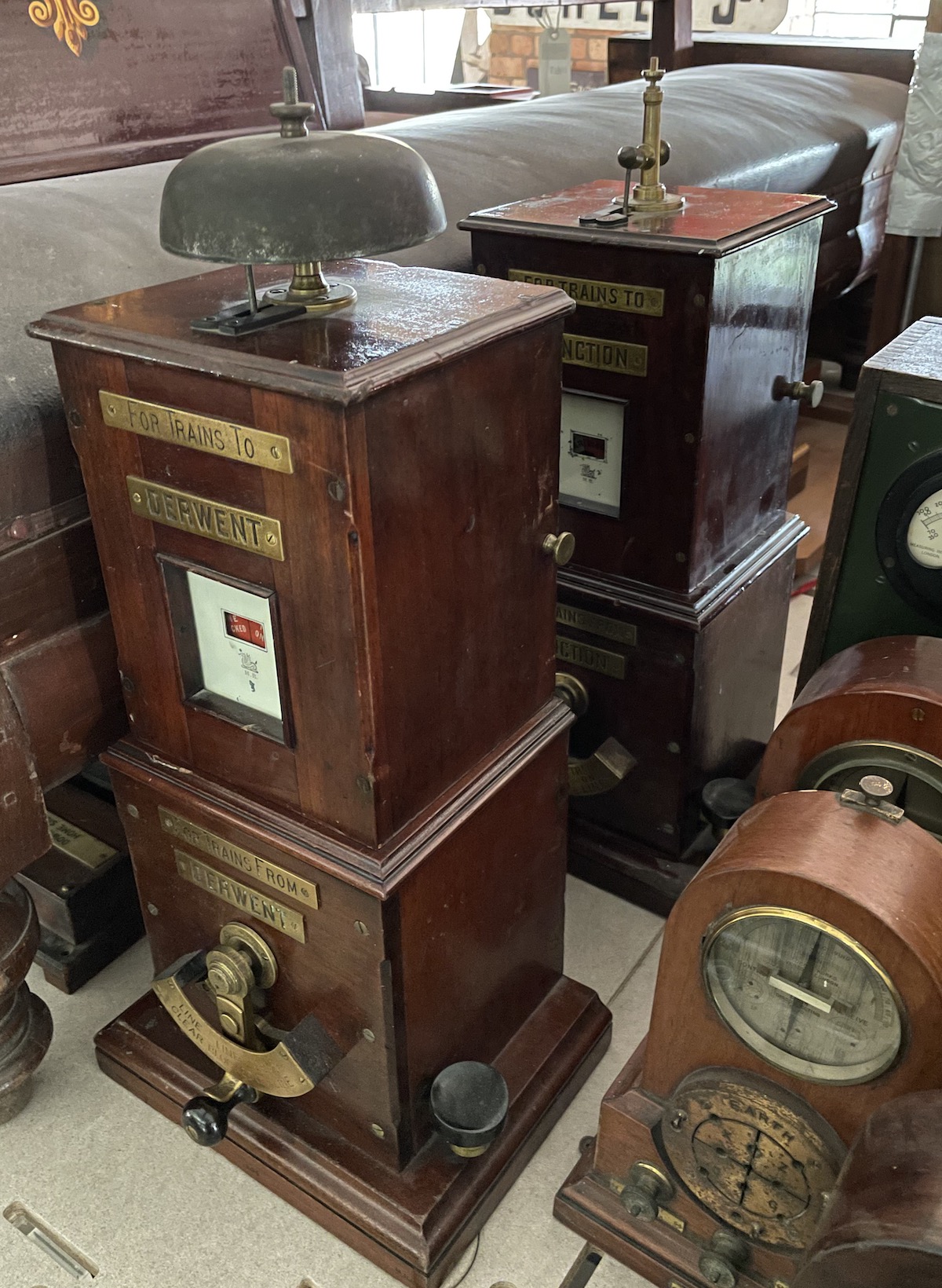 A pair of tall mahogany cased instruments with brass fittings. Both should have a domed bell on top but the one at the back is missing, revealing its striker. The front instrument has four brass plaques which combine to read For Trains To Derwent and For Trains From Derwent. There is a bell 'tapper' on the side.